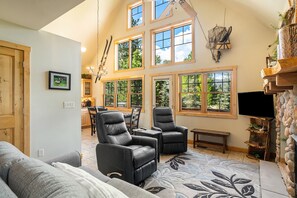 Living room with 2 leather recliners, sofa that seats 3, fireplace, flat screen TV, vaulted ceiling, and windows with view of outdoors.