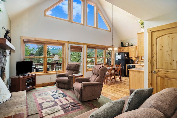 Open concept living room, with 2 reclining chairs, end table, flat screen TV, and lots of windows with view of mountains.