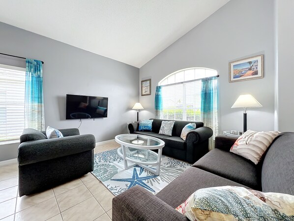 - Relax in a light filled living room with cozy seating and nautical themed decor
- Large windows and vibrant blue curtains create a fresh, coastal vibe throughout
- Watch your favorite shows on the wall mounted smart TV in this welcoming lounge space