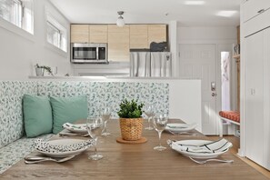 A modern dining area with a wooden table, green cushioned seats, and a potted plant centerpiece. The adjacent kitchen features stainless steel appliances and light wood cabinetry.