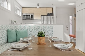 A modern dining area with a wooden table, green cushioned seats, and a potted plant centerpiece. The adjacent kitchen features stainless steel appliances and light wood cabinetry.
