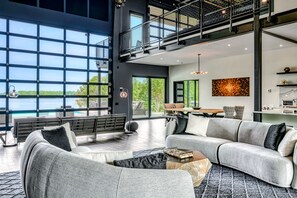 This image displays a modern, open-plan living space with an industrial ceiling, a sleek kitchen, and floor-to-ceiling windows that reveal a lake view.