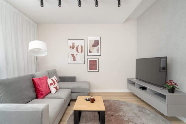  modern living room with a gray sofa, red and white accent pillows, a light wood coffee table, and wall art featuring abstract designs. There’s also a TV on a floating stand.