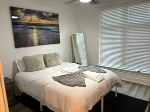 Serene bedroom with soft bedding, beach sunset artwork, and large window blinds.