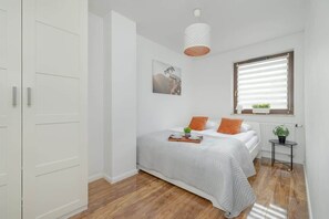 A cozy bedroom with a double bed, white walls, and a wooden floor. Orange pillows and a small bedside lamp add warmth to the decor.

