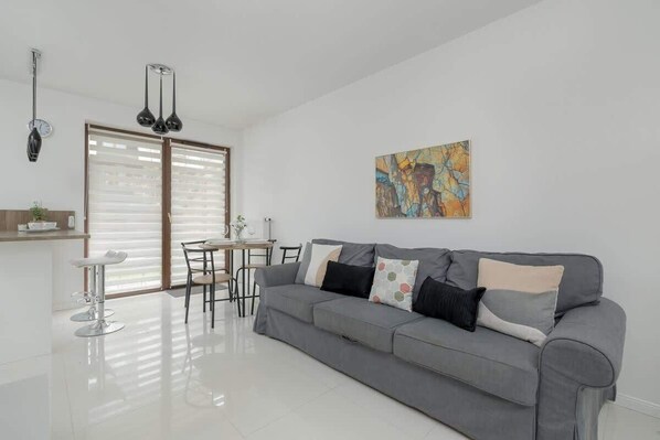 A bright living room with a gray sofa, a small round table, and large glass doors leading to the terrace. The space is minimalistic and modern.

