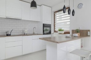 A sleek white kitchen with modern cabinets, a tiled backsplash, and a compact breakfast bar with two stools.

