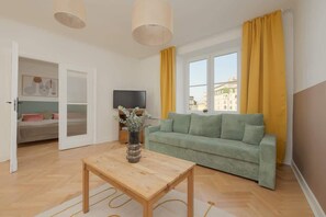 A bright living room with a green sofa, a glass coffee table, and natural light streaming in through large windows.

