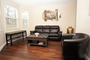 The living room is furnished with a dark leather sofa and armchair, a wooden coffee table, and a console against a window. An abstract wooden wall art piece adds a focal point, while the wooden floors and natural light create an inviting atmosphere.