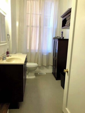 A well-appointed bathroom featuring a toilet, sink, and a mirror that adds a touch of elegance, with a shower stall adorned by a gracefully curtained window that bathes the space in gentle natural light.