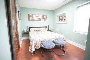 The bedroom exudes a serene atmosphere with its soft green walls, accented by coordinating artwork. A floral bedspread graces the bed, and two unique metal stools sit at the foot, adjacent to the rustic sliding barn door.