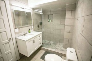The bathroom boasts a sleek design with gray tile work throughout, a spacious walk-in shower, and a vanity with ample storage. The mirror above the sink is well-lit, creating a bright and functional space.
