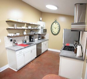 In a warm, inviting kitchen, white cabinetry and stainless steel appliances contrast with terracotta-colored floors. Open shelves display dishware neatly, while a large wall clock adds a touch of classic charm.