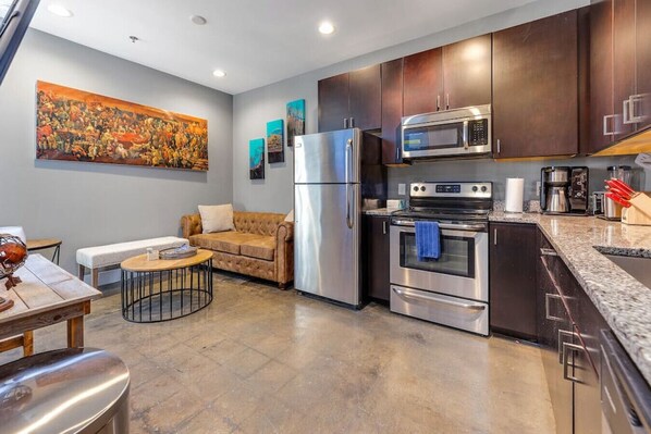 The modern kitchen is equipped with stainless steel appliances, dark wood cabinets, and granite countertops. The space is well-organized with a coffee maker and other essentials, complemented by under-cabinet lighting.