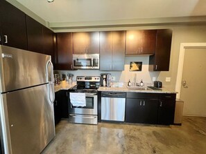 The kitchen is sleek with modern stainless steel appliances, rich dark cabinetry, and granite countertops. It is well-equipped and designed for functionality, with polished concrete floors adding an industrial touch.