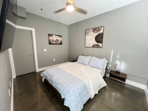 The bedroom is modern and bright with a comfortable bed, flanked by nightstands and decorative lamps. Large artworks add a splash of color, and a ceiling fan provides comfort. A wall-mounted TV offers entertainment.