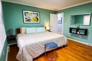 The bedroom is serene and stylish, with teal walls and polished hardwood floors. Coordinating pillows on the bed complement the abstract wall art. A modern stool and a floating shelf add functional elegance to the space, while natural light enhances the r