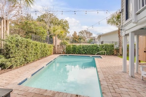 A rectangular outdoor pool with clear water surrounded by brick paving, greenery, and string lights overhead.