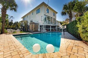 A modern two-story house with a backyard pool, surrounded by patio furniture and palm trees, under a clear blue sky.