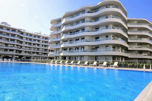 A luxurious modern apartment complex with curved balconies and a large swimming pool reflecting the contemporary architecture.

