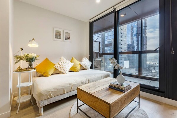 A living area sits alongside floor-to-ceiling glass windows, and has light-washed timber floors, a sofa, timber coffee table and a flat-screen TV.
