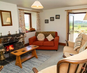 Living room with open fire, sofa, armchairs, coffee table, doors open to garden