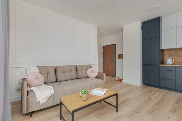 A spacious living room with a neutral-toned sofa and a coffee table, complemented by light wood flooring. The open-plan design features a modern kitchen in the background with dark and white cabinetry.