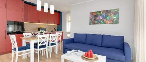Cozy living room with blue sofa, white coffee table, dining area in background, and bright artwork on wall.
