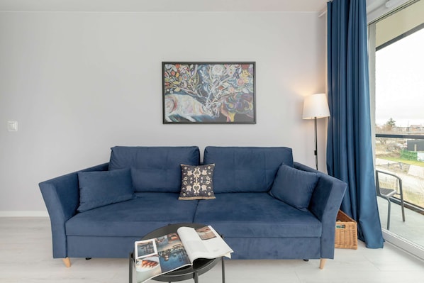 A spacious living area with a blue sofa, dining table, and large windows offering plenty of sunlight.