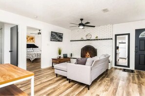 The living room is a cozy and elegant space, featuring a fireplace that adds warmth, a wall mirror, natural light, a comfortable sofa, a table, décor, a TV, and a ceiling fan.