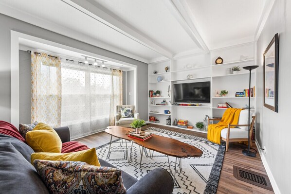 Bright living space with a plush sofa and accent chairs arranged around a live-edge coffee table on a patterned rug. Built-in white shelves flank a wall-mounted TV, while sheer curtains filter soft natural light through large windows. 🛋️📺🌿