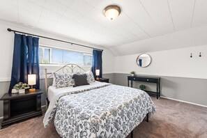 Cozy bedroom features a metal-framed bed dressed in blue-and-white floral linens, flanked by matching dark nightstands with warm bedside lamps. A long transom window above is framed by navy curtains, letting in natural light. The lower half of the walls i