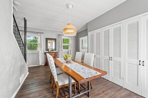 Bright dining area showcases a live-edge wooden table with a lace runner, surrounded by white upholstered chairs and topped with a bowl of green apples. A woven pendant light hangs above, while a row of white louvered closet doors lines the gray wall. Sun