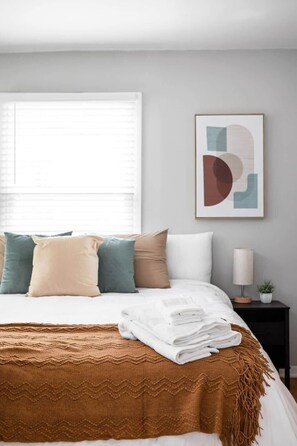 Our snug bedroom features comfortable beds adorned with fresh bed sheets and blankets, plush pillows, and fluffy towels. Enjoy the natural light streaming in through the window, along with the convenience of a bedside table and lamp for your nighttime reading. Admire the thoughtful wall decor that adds charm to the room.