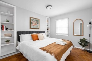 Welcome to our inviting bedroom, complete with crisp, fresh linens and plush pillows to ensure your comfort. Bask in the natural light pouring in through the window, accompanied by the soft glow of a stylish lamp. Take pleasure in the plant decor, wall frame, and utilize the mirror and shelves or organizer on both sides for added convenience and charm.