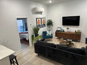 Cozy and stylish living space featuring a plush black sofa, chic glass coffee table, and a flat-screen TV, complemented by natural light and tasteful decor.