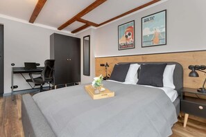 A bright double bedroom with a soft gray bedspread, wooden headboard, and framed vintage-style posters.