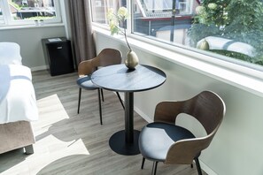 This bright and airy corner features a round black table and two modern chairs with wooden accents, set next to large windows offering a view of the neighborhood and greenery. Perfect for enjoying a meal, working, or simply relaxing with plenty of natural light.