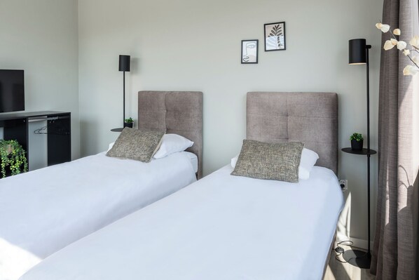 This bright bedroom features two comfortable single beds with plush headboards and decorative pillows, ideal for a relaxing stay. Each bed has its own modern bedside lamp and table with a small plant. Minimal wall decor and natural light create a serene and inviting atmosphere.