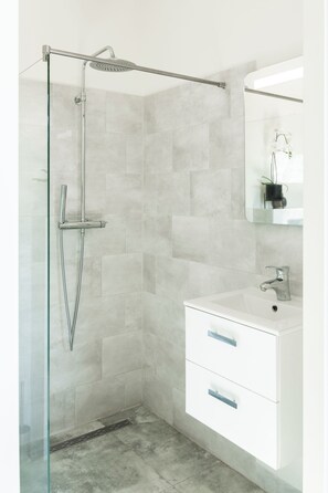 A sleek and modern bathroom featuring a walk-in rain shower with a glass partition and light gray tiled walls. The space includes a white vanity with two drawers for storage and a mounted mirror above. Bright and minimalist, ideal for a refreshing experience.