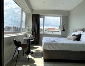 This bright and modern bedroom features a large window providing plenty of natural light and offering a rooftop view of the surroundings. It includes a comfortable double bed, a small round table with two chairs, and minimalist decor in neutral tones. Perfect for a relaxing stay.
