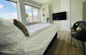 This cozy bedroom offers a modern design with a comfortable bed and plush cushions. The room features a wall-mounted TV and a stylish shelving unit, accented with natural light from large windows and minimalist decor for a serene ambiance.