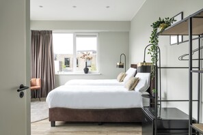 Bright and modern bedroom with twin beds, white linens, gold-accented lamps, greenery, open shelving, neutral tones, and a large window for natural light.