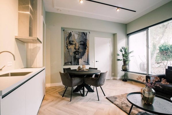 A chic and modern living space featuring a dining area with comfortable chairs, a beautiful piece of art on the wall, and a window that opens up to a lush garden area, creating a relaxing atmosphere.