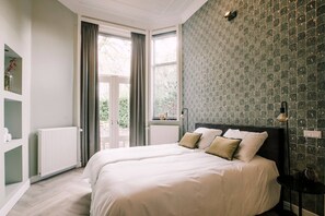 A cozy bedroom with soft, neutral-toned bedding and a sophisticated feature wall in calming shades of green. Large windows let in natural light, creating a serene and inviting space to relax.