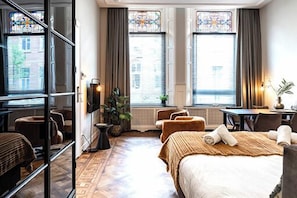 A cozy, inviting bedroom featuring two large windows with city views, offering a serene retreat with plush bedding and a warm, ambient glow.