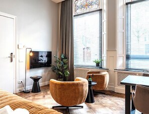 Bright and elegant living area with large stained-glass windows, cozy velvet chairs, a wall-mounted TV, and warm wood flooring that adds a touch of charm.