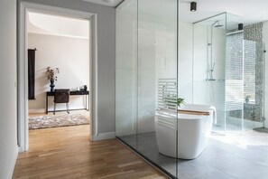 A bright, elegant space featuring a glass-enclosed bathroom with a freestanding tub, rainfall shower, and a cozy desk area in the adjacent room.