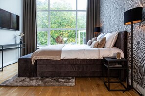 
A luxurious bedroom with a plush bed, zebra-patterned wallpaper, floor-to-ceiling windows, soft lighting, cozy accents, and a sleek console table with decor.