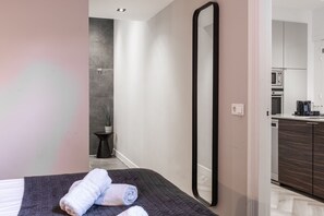 A serene bedroom view featuring a neatly made bed with rolled towels, soft lighting, and a full-length mirror. The hallway leads to a modern kitchen and sleek bathroom.
